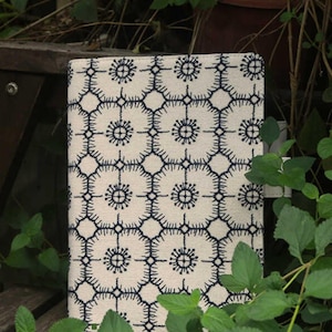 Op de afbeelding: Een witte stoffen notitieboekomslag met een blauw geometrisch patroon. Het patroon heeft herhalende cirkels met een stervormig ontwerp.