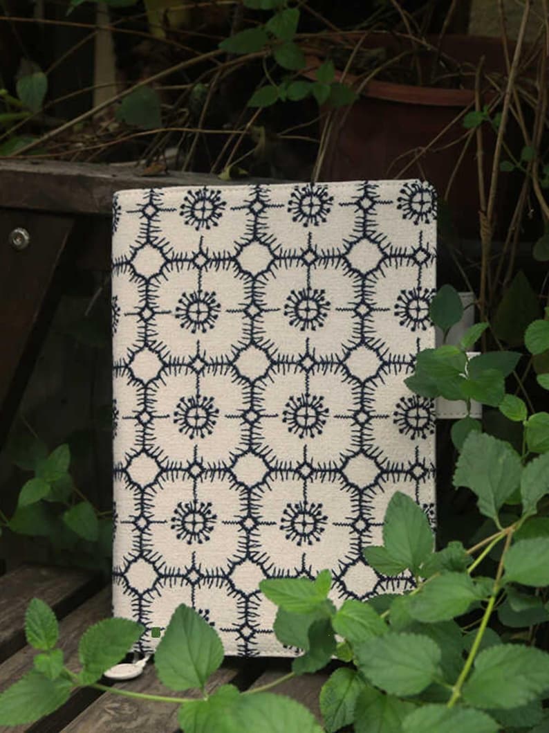 Op de afbeelding: Een witte stoffen notitieboekomslag met een blauw geometrisch patroon. Het patroon heeft herhalende cirkels met een stervormig ontwerp.