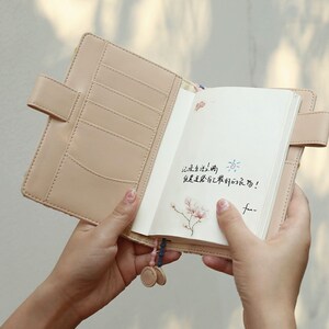 May include: A light brown leather bound journal with a floral design on the cover. The journal is open to a blank page with a handwritten message in black ink that says "记录生活的小确幸, 继续走下去, 有期待的活著!" and a small flower illustration. The journal has a pink and blue beaded tassel.