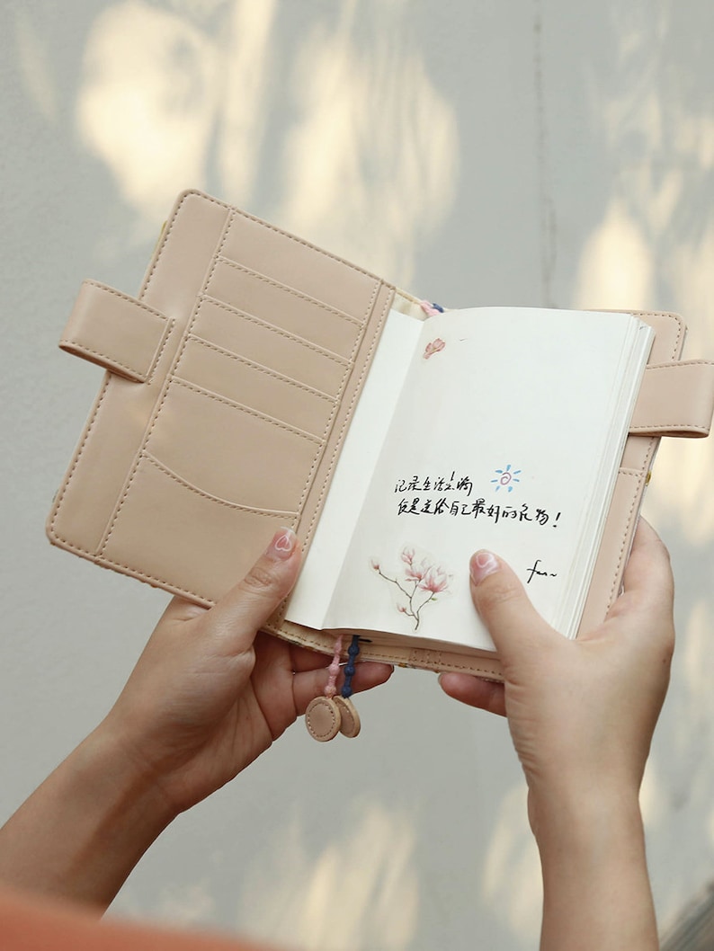 May include: A light brown leather bound journal with a floral design on the cover. The journal is open to a blank page with a handwritten message in black ink that says "记录生活的小确幸, 继续走下去, 有期待的活著!" and a small flower illustration. The journal has a pink and blue beaded tassel.