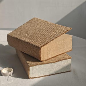 May include: Two decorative books with beige burlap covers. One book is open, revealing the pages. The other book is closed. The books are stacked on top of each other. A roll of tape and a small piece of paper are in the lower left corner.