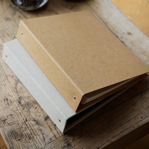 Puede incluir: Dos carpetas de anillas, una gris claro y otra marrón claro, apiladas sobre una superficie de madera. Las carpetas tienen un diseño sencillo con remaches metálicos a lo largo del lomo. La carpeta marrón claro está encima.