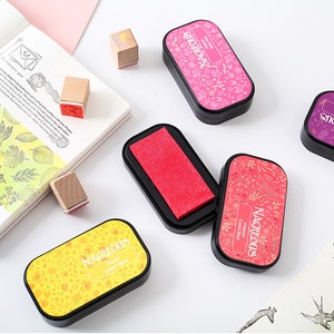 May include: Four ink pads with floral designs and the word "Nacreous" printed on the top. The ink pads are in the colors coral red, lemon yellow, red, and pink.