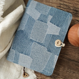 Op de afbeelding: Een blauwe denim notitieboekomslag met een patchwork ontwerp. De omslag heeft een knoopsluiting en een bruine leren kwast.