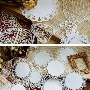 May include: A set of white paper doilies with intricate lace designs. The doilies are arranged on a book with a brown cover and a white background. The book has text in a foreign language. The doilies are perfect for adding a touch of elegance to any occasion.