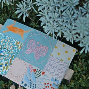 May include: A blue fabric patchwork notebook cover with a pink butterfly embroidery, a white circle, and a floral design. The cover is decorated with small white beads.