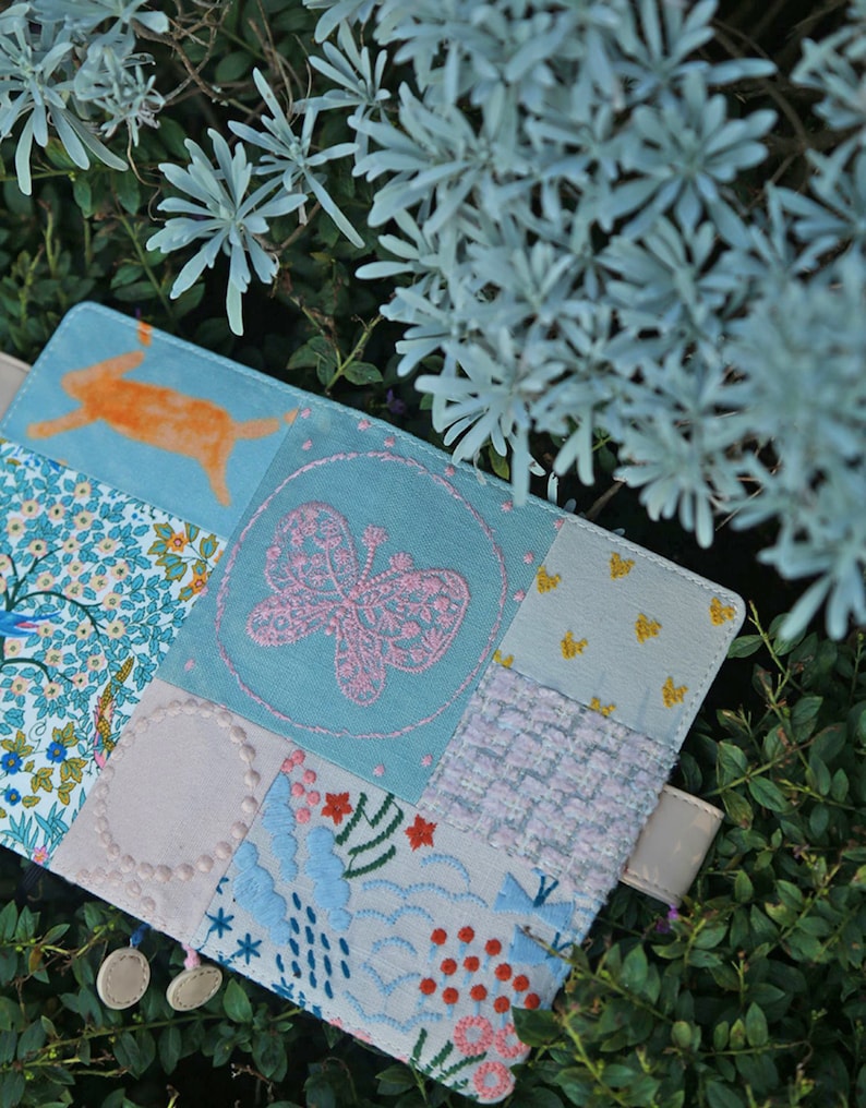 May include: A blue fabric patchwork notebook cover with a pink butterfly embroidery, a white circle, and a floral design. The cover is decorated with small white beads.