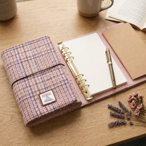 May include: A pink and purple plaid wool planner cover with a Harris Tweed label, featuring a gold six-ring binder mechanism, blank white paper, a brown notebook insert, and a metallic pen. The cover is secured with a thin dark brown elastic band.