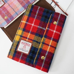 Pode incluir: Uma capa de caderno Harris Tweed com um padrão xadrez vermelho, azul, amarelo e branco arrojado. Possui um fecho de pressão de latão e uma etiqueta com o logótipo Harris Tweed. Um segundo caderno com um padrão xadrez rosa e azul também é visível.