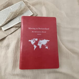 May include: A red journal with the text "Moving to Switzerland, My Adventures Abroad, 2026" and a white world map graphic. The journal is lying on a cream-colored fabric.