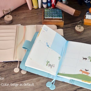 Puede incluir: Dos cuadernos abiertos con cubiertas de cuero. Uno es beige y el otro es azul. Ambos cuadernos tienen un marcador de cinta y un bolsillo en el interior de la cubierta. El cuaderno beige tiene un dise&ntilde;o de calendario y el cuaderno azul tiene una p&aacute;gina en blanco.