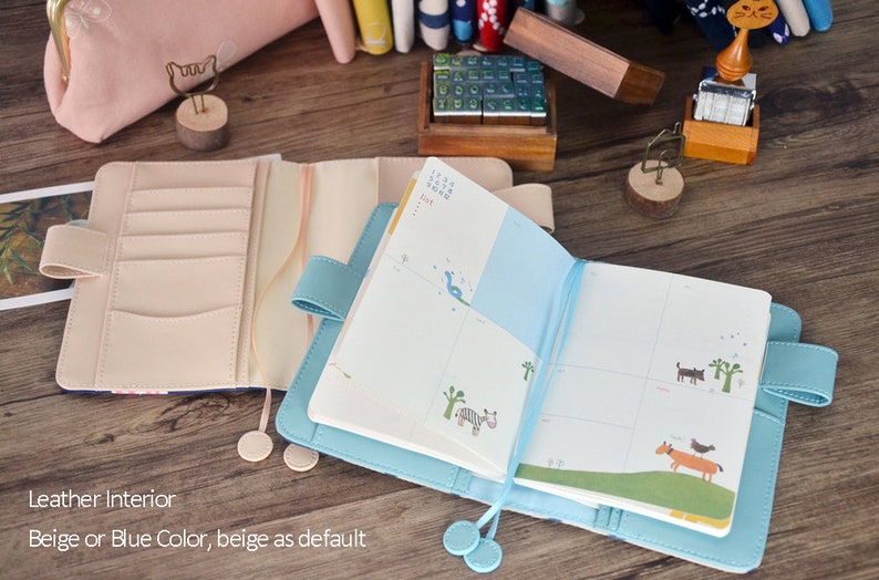 Puede incluir: Dos cuadernos abiertos con cubiertas de cuero. Uno es beige y el otro es azul. Ambos cuadernos tienen un marcador de cinta y un bolsillo en el interior de la cubierta. El cuaderno beige tiene un dise&ntilde;o de calendario y el cuaderno azul tiene una p&aacute;gina en blanco.