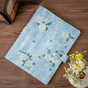 May include: A light blue journal with white floral embroidery and green leaves. The journal has a button closure and is decorated with small, scattered beads. The cover has a textured appearance.