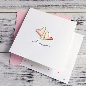Puede incluir: Una tarjeta blanca con un diseño de corazón de acuarela rosa y blanco y la palabra "love" escrita en tinta negra. La tarjeta está rodeada por un sobre rosa.