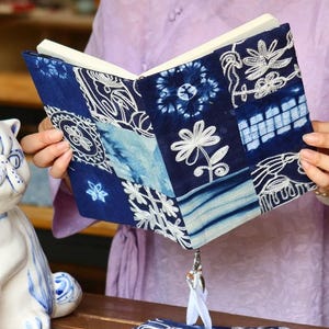 Könnte beinhalten: Ein gebundenes Buch mit einem Stoffeinband im Patchwork-Design aus dunkelblauen und hellblauen Indigofarben. Der Einband zeigt weiße, gestickte Blumen- und Geometriemuster sowie ein weißes Lesezeichenband mit einem silbernen Metallanhänger.