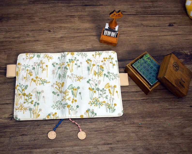 May include: A white fabric notebook cover with a floral pattern in shades of green and yellow. The cover has two leather straps and two leather tassels. The notebook is open and the pages are blank.