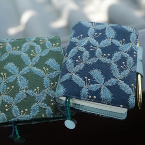 May include: Two notebooks with embroidered floral patterns. One is navy blue with light blue embroidery, the other is green with light blue embroidery. A gold pen is inserted into the blue notebook. Both have matching bookmarks.