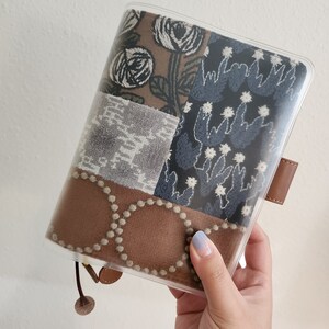 May include: A clear plastic cover for a planner or notebook. The cover has a brown, gray, and blue patchwork design with floral and geometric patterns. The cover has a brown leather strap with a metal buckle.
