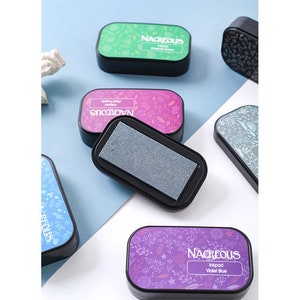 May include: A set of six ink pads with floral designs and the word "Nacreous" printed on each. The ink pads are in various colors, including green, pink, blue, and purple. The ink pads are labeled with the ink color, such as "Emerald Green" and "Violet Blue".