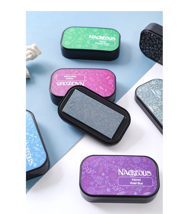 May include: A set of six ink pads with floral designs and the word "Nacreous" printed on each. The ink pads are in various colors, including green, pink, blue, and purple. The ink pads are labeled with the ink color, such as "Emerald Green" and "Violet Blue".