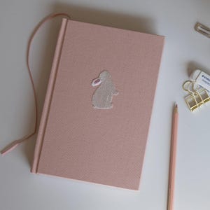 Può includere: Un taccuino rivestito in tessuto rosa chiaro con un piccolo coniglio bianco ricamato che presenta un dettaglio rosa all'interno dell'orecchio. Il taccuino include un segnalibro a nastro rosa abbinato, una matita rosa chiaro e una clip dorata.