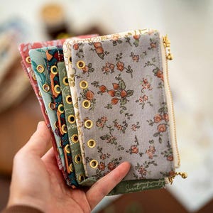 以下が含まれることがあります： グレー、赤、緑など、さまざまな色の花柄の生地ノートの束。各ノートには金色のハトメとジッパーが付いています。ノートは手で持たれています。