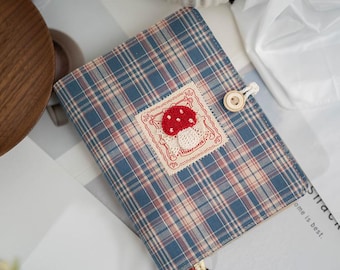 May include: A blue and red plaid fabric journal cover with a red mushroom applique. The applique is on a white lace patch with red trim. The journal cover has a wooden button closure.