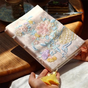 Può includere: Un diario di seta rosa con un ricamo floreale. Il diario ha un fiocco e è chiuso.