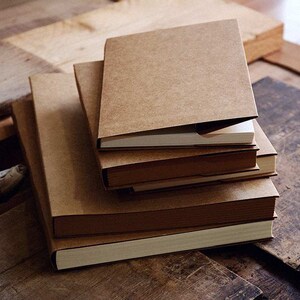 May include: A stack of four brown notebooks with blank white pages. The notebooks are bound with a brown cover and have a rustic, vintage aesthetic.