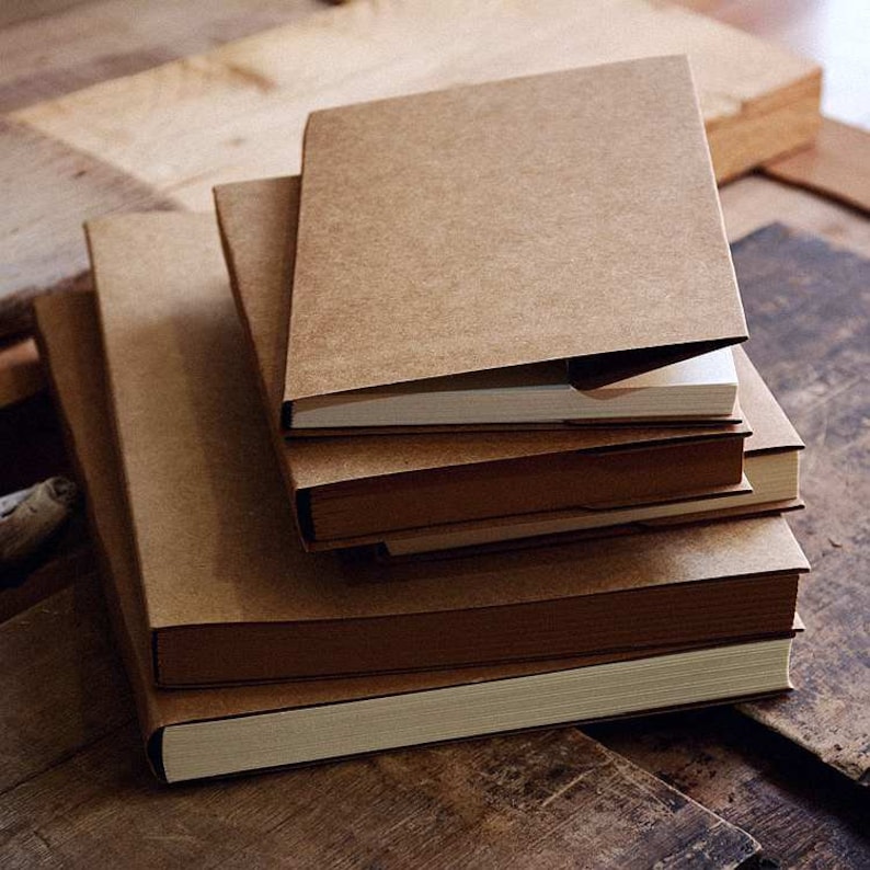 May include: A stack of four brown notebooks with blank white pages. The notebooks are bound with a brown cover and have a rustic, vintage aesthetic.
