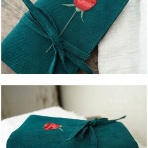 May include: A green corduroy notebook with a red embroidered rose and a green ribbon tied around it. The notebook has two brass rings on the side.