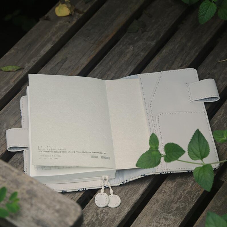 Op de afbeelding: Een wit notitieboek met een grijze omslag en een bloemenpatroon. Het notitieboek is open op een lege pagina en heeft een lint-bladwijzer. Het notitieboek ligt op een houten oppervlak.