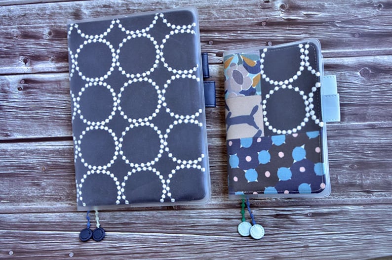 May include: Two fabric-covered notebooks with clear plastic covers. The larger notebook has a dark blue cover with a white circle pattern. The smaller notebook has a patchwork cover with blue, brown, and white patterns.