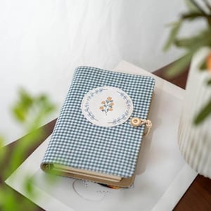 May include: A small notebook with a blue and white gingham pattern cover. A round patch with a floral design is centered on the cover. The notebook is closed with a button and loop closure. The notebook is resting on a white surface.