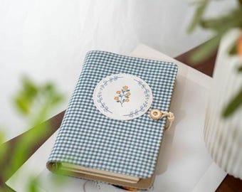 May include: A small notebook with a blue and white gingham pattern cover. A round patch with a floral design is centered on the cover. The notebook is closed with a button and loop closure. The notebook is resting on a white surface.
