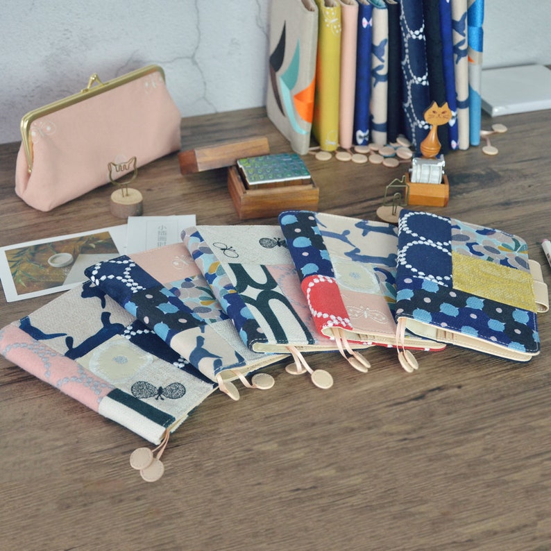 Puede incluir: Seis cuadernos de tela con diferentes patrones y dise&ntilde;os. Los cuadernos est&aacute;n dispuestos en una fila sobre una mesa de madera. Cada cuaderno tiene un marcap&aacute;ginas de tela.