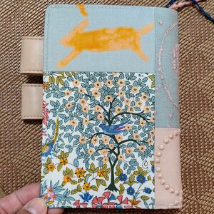 May include: A light blue fabric notebook cover with a patchwork design. The cover features a yellow rabbit, a floral pattern, and a pink fabric with embroidered dots. The cover has two beige leather straps.