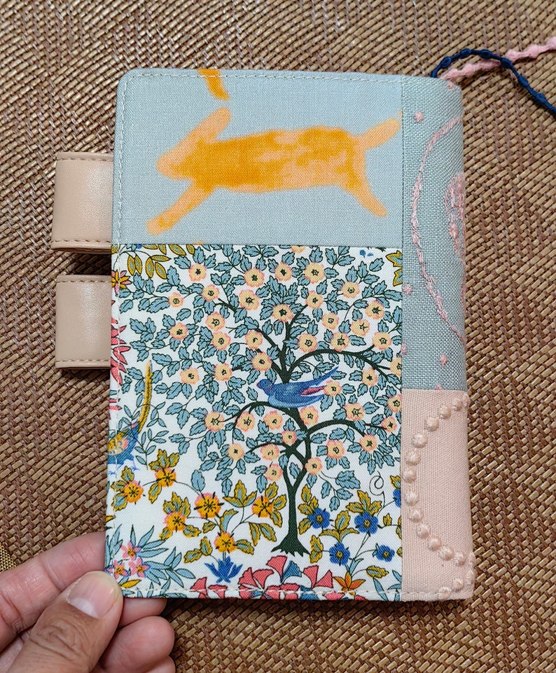May include: A light blue fabric notebook cover with a patchwork design. The cover features a yellow rabbit, a floral pattern, and a pink fabric with embroidered dots. The cover has two beige leather straps.