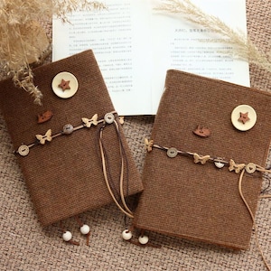 May include: Two rectangular brown fabric-covered journals featuring a textured weave. Each journal is decorated with a large cream button with a star cutout, a small wooden rocking horse, and a cord wrapped around with wooden butterfly and circular buttons.