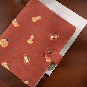 Puede incluir: Un archivador de tela roja con un patrón de calabaza bordado en la parte delantera. El archivador tiene una correa de cuero marrón con un cierre de botón de latón. El texto "WOOL STITCH" está impreso en el papel blanco dentro del archivador.