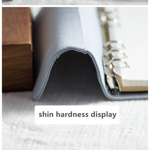 May include: A pale blue journal featuring floral embroidery in orange, brown, and teal. It has a matching cord closure with a pearl and a tassel. The text "shin hardness display" is visible.