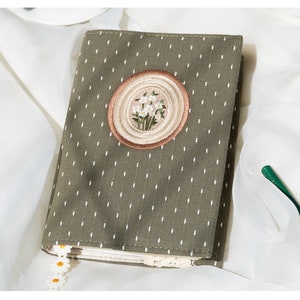 May include: A green fabric notebook cover with a white daisy embroidery design in a brown circle. The cover has a white polka dot pattern.