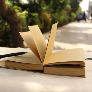 May include: A brown leather-bound journal with a black pen lying on top of it. The journal is open and the pages are blank. The journal is sitting on a white surface.