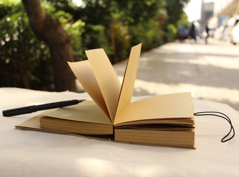 May include: A brown leather-bound journal with a black pen lying on top of it. The journal is open and the pages are blank. The journal is sitting on a white surface.