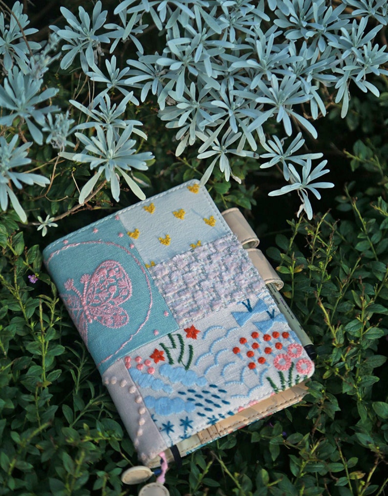 May include: A small, blue and white patchwork notebook with a butterfly design and floral embroidery. The notebook is lying on a bed of green leaves.