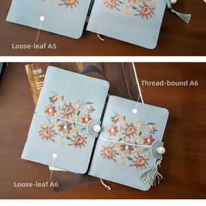 May include: Pale blue notebooks with embroidered floral patterns in orange, brown, and teal. Available in A5 and A6 sizes, with loose-leaf and thread-bound bindings. Each notebook has a pearl clasp and a tassel for closure.