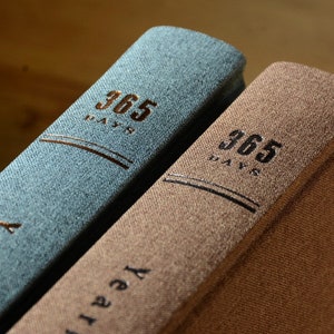 May include: Two hardcover journals with textured covers. The journal on the left is blue and the journal on the right is brown. Both journals have the text "365 Days" printed on the front cover.