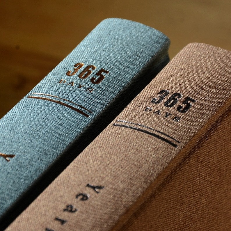 May include: Two hardcover journals with textured covers. The journal on the left is blue and the journal on the right is brown. Both journals have the text "365 Days" printed on the front cover.