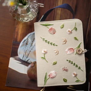 May include: A beige linen notebook cover with embroidered pink tulips, white flowers, and green leaves. The cover has a button closure and a blue strap.