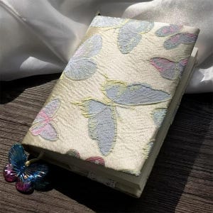 Pode incluir: Uma capa de caderno de cor creme com um delicado padrão de borboletas em tons de rosa, azul e amarelo. A capa é feita de um tecido texturizado e tem um brilho sutil.